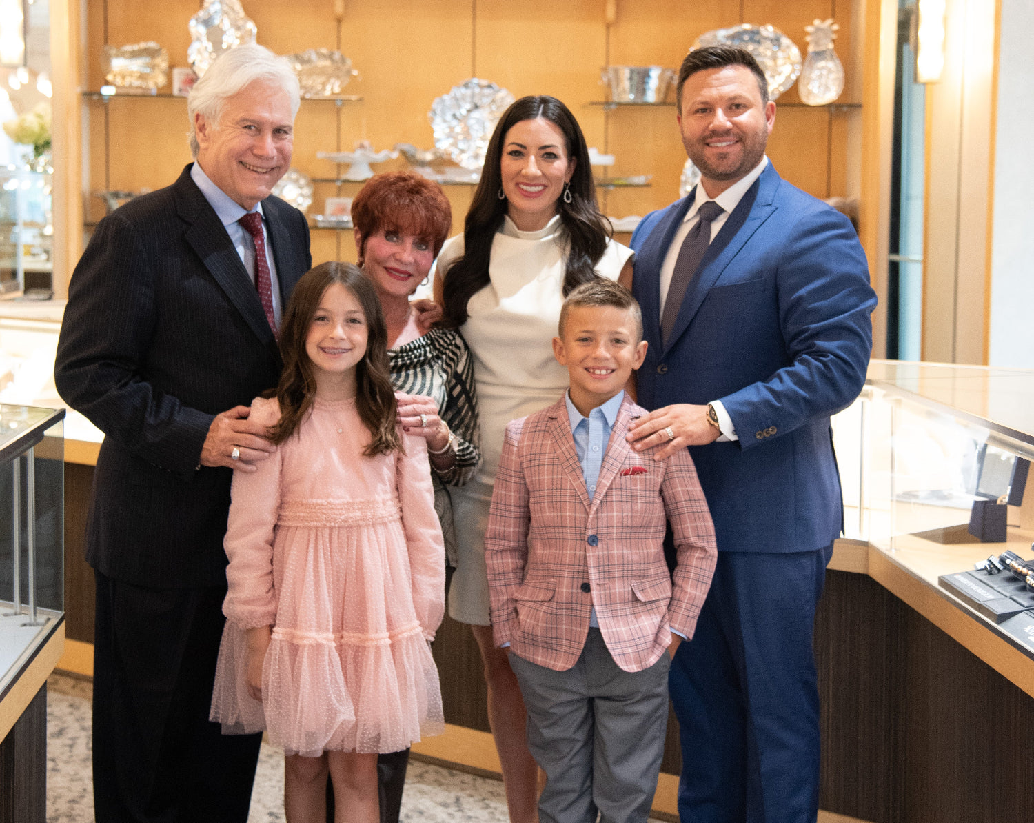 A family of five posing together inside a luxury jewelry showroom. The group includes a man in a black suit, a woman in a cream top, a man in a blue suit, a young boy in a pink plaid blazer, a young girl in a pink tulle dress, and a woman in a black and white patterned top.